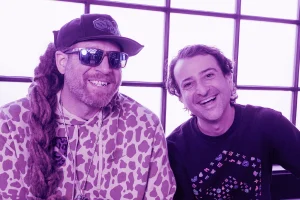 Two men smiling indoors in front of large windows, including one wearing sunglasses and a patterned hoodie, in a cannabis-themed lounge with purple lighting.